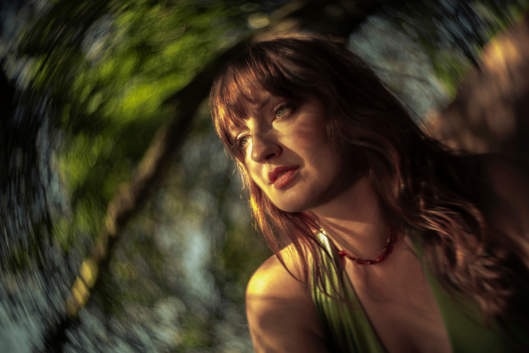 Madison Cunningham looks to the right of the camera, framed by a swirling effect behind the lens. She appears to be in the forest, and has sunlight across her face.